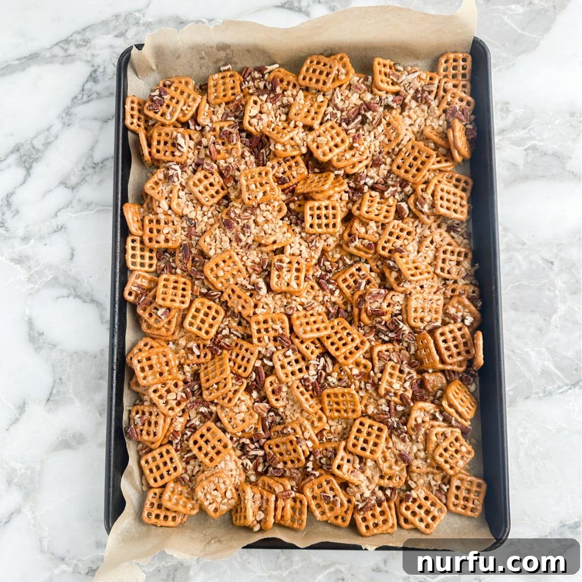 A baking pan filled with golden-brown butter toffee pretzels, glistening with the caramel glaze and generously studded with toffee bits and chopped pecans, fresh out of the oven.