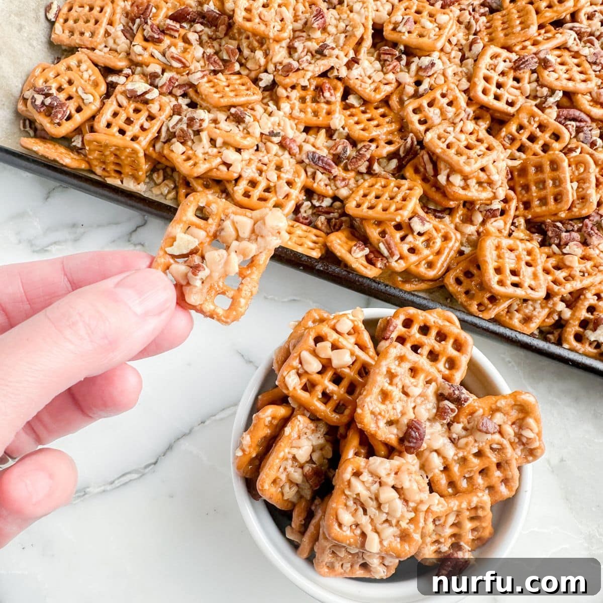 A hand gently holding a single butter toffee pretzel, showcasing its golden glaze, visible toffee bits, and crunchy texture against a soft, blurred background, ready to be enjoyed.