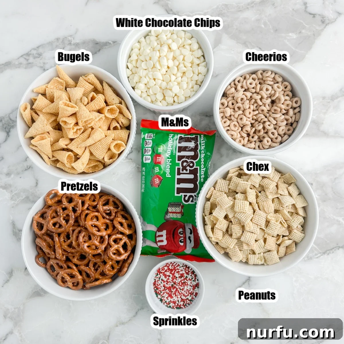 Festive Reindeer Treats 3 A vibrant display of key Christmas reindeer crack recipe ingredients laid out in separate bowls, including white chocolate chips, Cheerios, various pretzels, Bugles, festive sprinkles, and Chex cereal, ready to be combined.