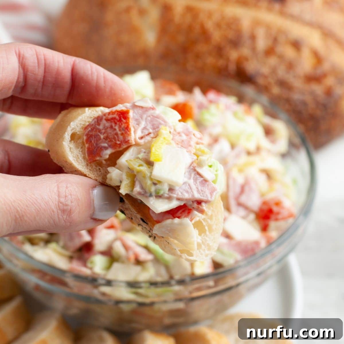 Stacked Hoagie Dip 7 A hand reaching in to grab a piece of toasted bread generously topped with the vibrant and chunky hoagie dip, ready to be enjoyed.