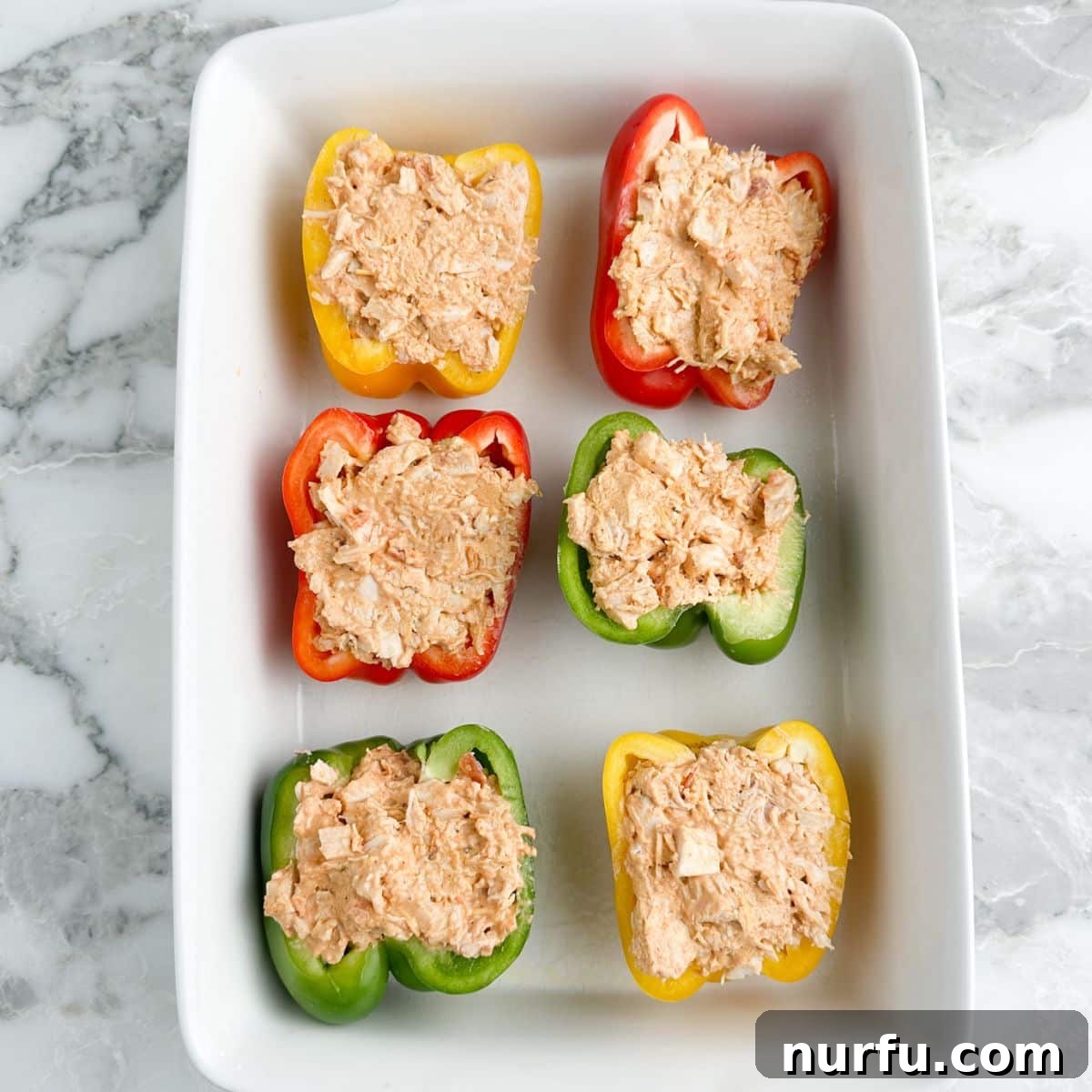 Halved bell peppers generously stuffed with a creamy chicken mixture, arranged in a baking dish before topping with cheese.