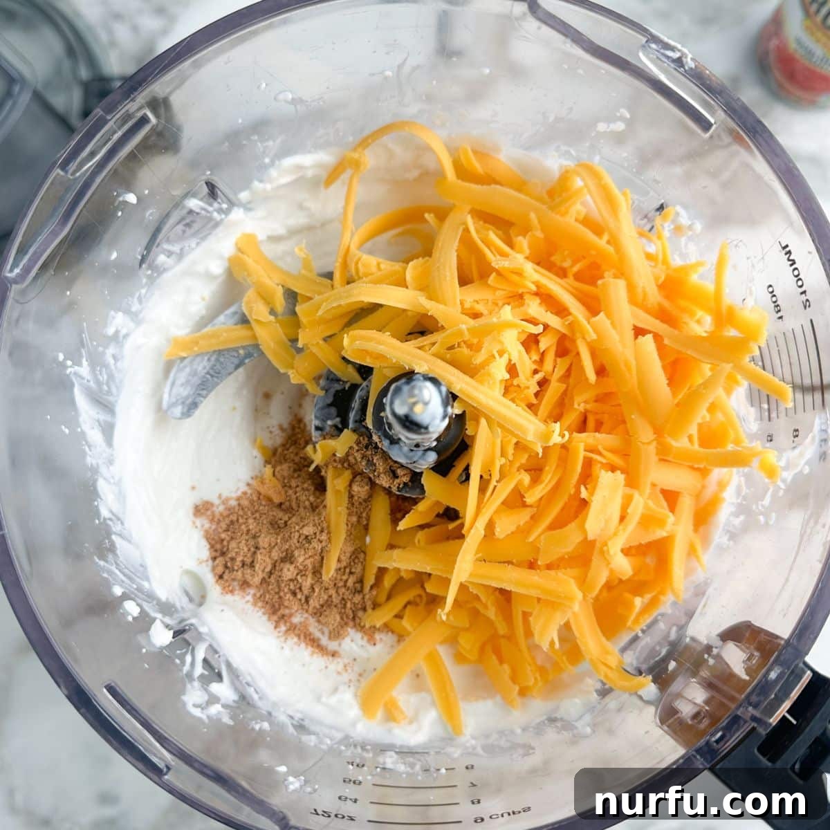 Inside a food processor, a blend of whipped cottage cheese, shredded cheese, and taco seasoning creates a rich, orange-hued mixture, illustrating the mid-preparation stage of the queso.