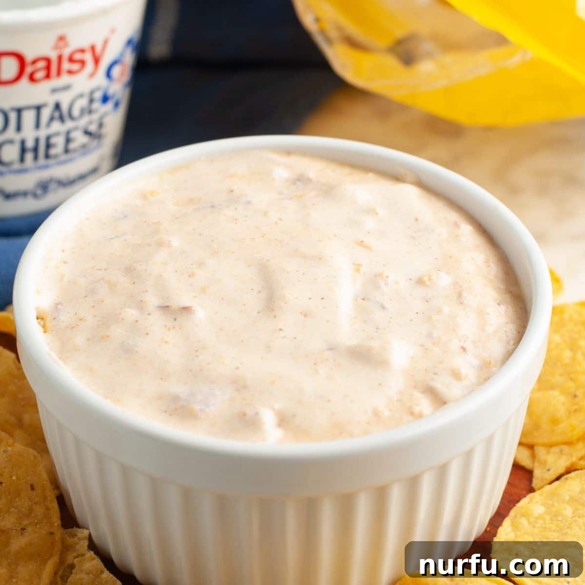 A generous serving of cottage cheese queso dip in a rustic bowl, perfectly smooth and adorned with a sprinkle of fresh herbs, ready for serving alongside an assortment of dippers. The composition highlights the dip's appealing texture and vibrant color.