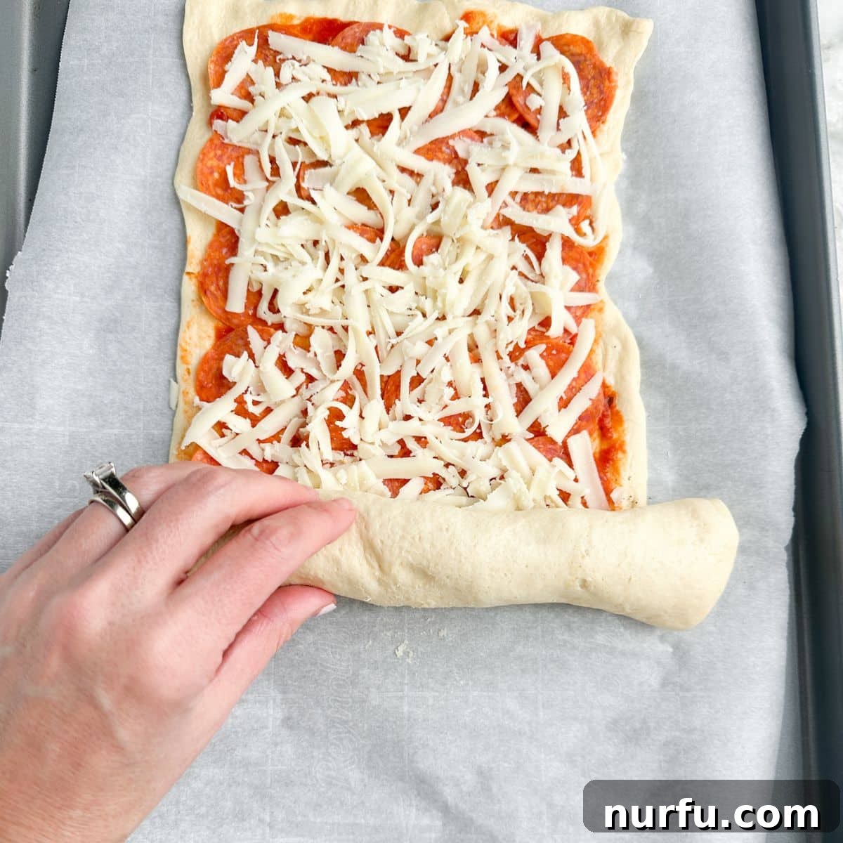 Pepperoni Pizza Swirls 8 Hands carefully rolling crescent dough, filled with pizza sauce and cheese, into a tight log on parchment paper.