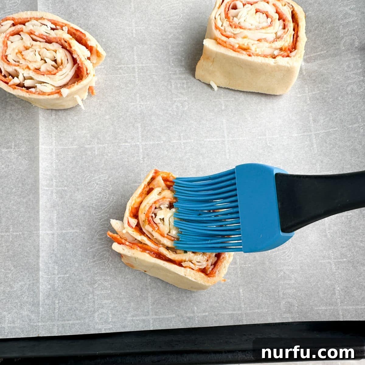 Pepperoni Pizza Swirls 10 Unbaked pizza pinwheels on a baking sheet, being brushed with a savory butter mixture, preparing them for a golden finish.