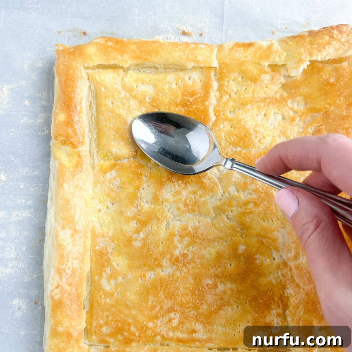 Quick Puff Pastry Pizza 7 A spoon gently pressing down the puffed-up center of a partially baked puff pastry, creating a well for toppings.