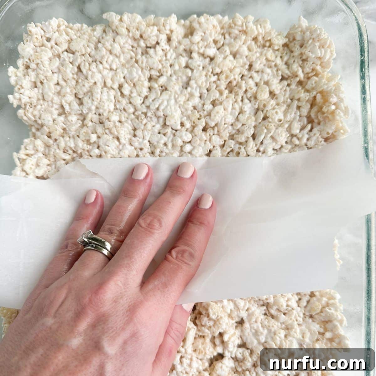 Cloud Nine Krispie Treats 6 A hand pressing down Rice Krispie treats into a prepared baking dish using a spatula.