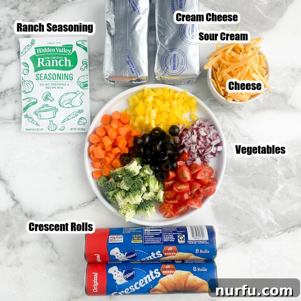 Flaky Garden Veggie Bake 3 Ingredients for crescent roll veggie pizza laid out: cream cheese, cans of crescent rolls, a plate of assorted chopped vegetables, a bowl of shredded cheese, and a packet of ranch seasoning.