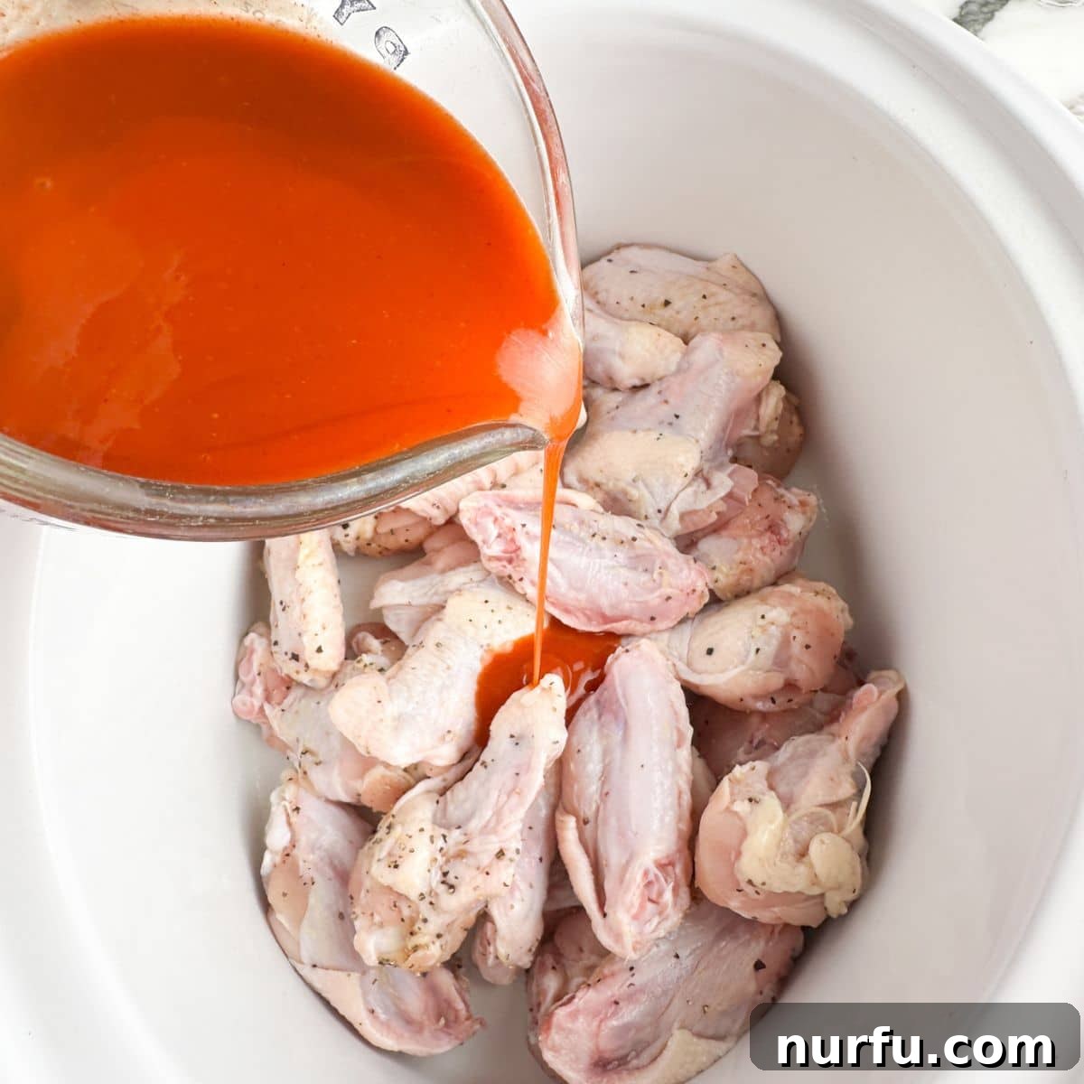 Effortless Slow Cooker Chicken Wings 6 Seasoned chicken wings arranged in a slow cooker, with buffalo sauce being poured over them.