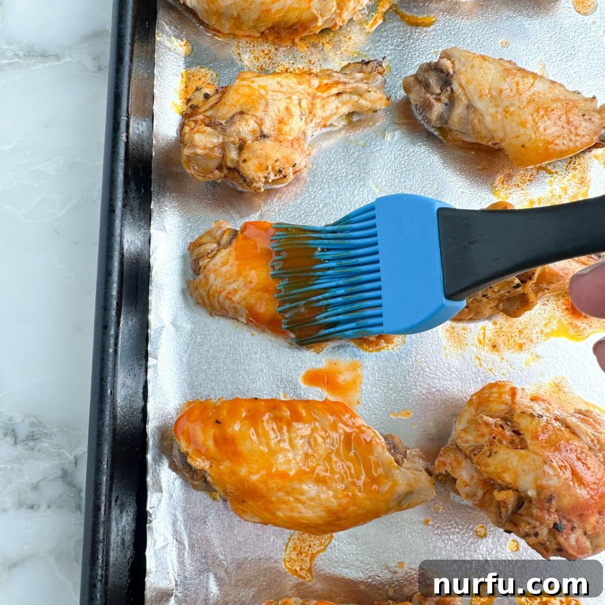 Effortless Slow Cooker Chicken Wings 9 A hand brushing additional buffalo sauce onto golden-brown, cooked chicken wings on a baking sheet before final broiling.