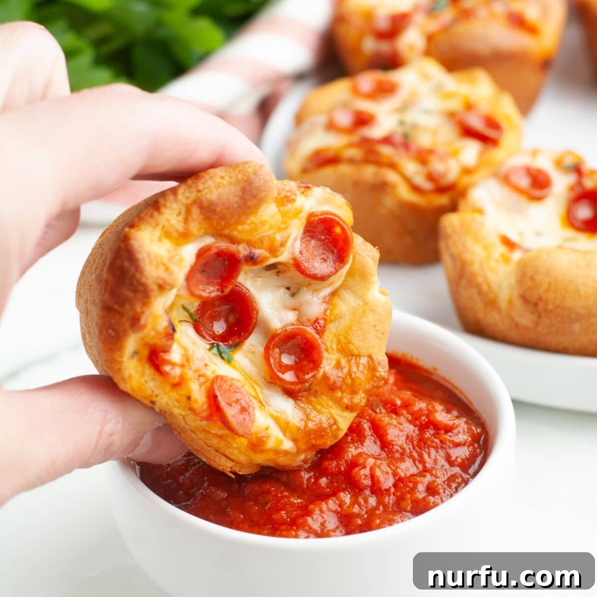 Golden brown pizza cupcake being dipped into a bowl of vibrant red marinara sauce, showcasing its cheesy topping and crisp crescent roll crust. 
