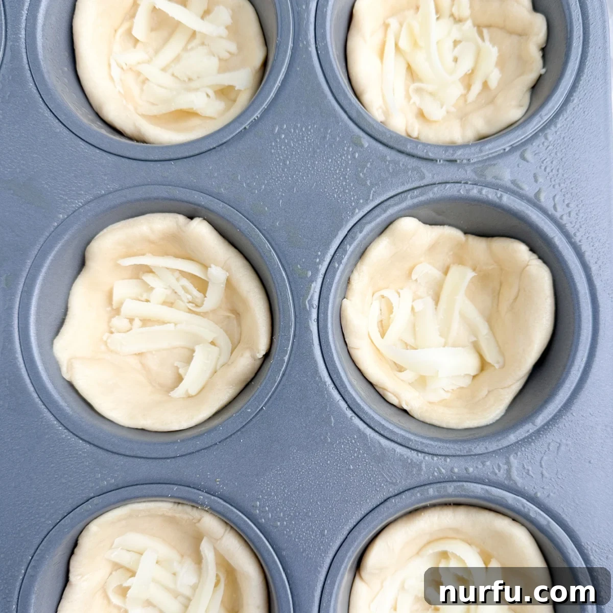 Muffin tin with crescent dough crusts, each lightly sprinkled with garlic powder and a layer of shredded mozzarella cheese.
