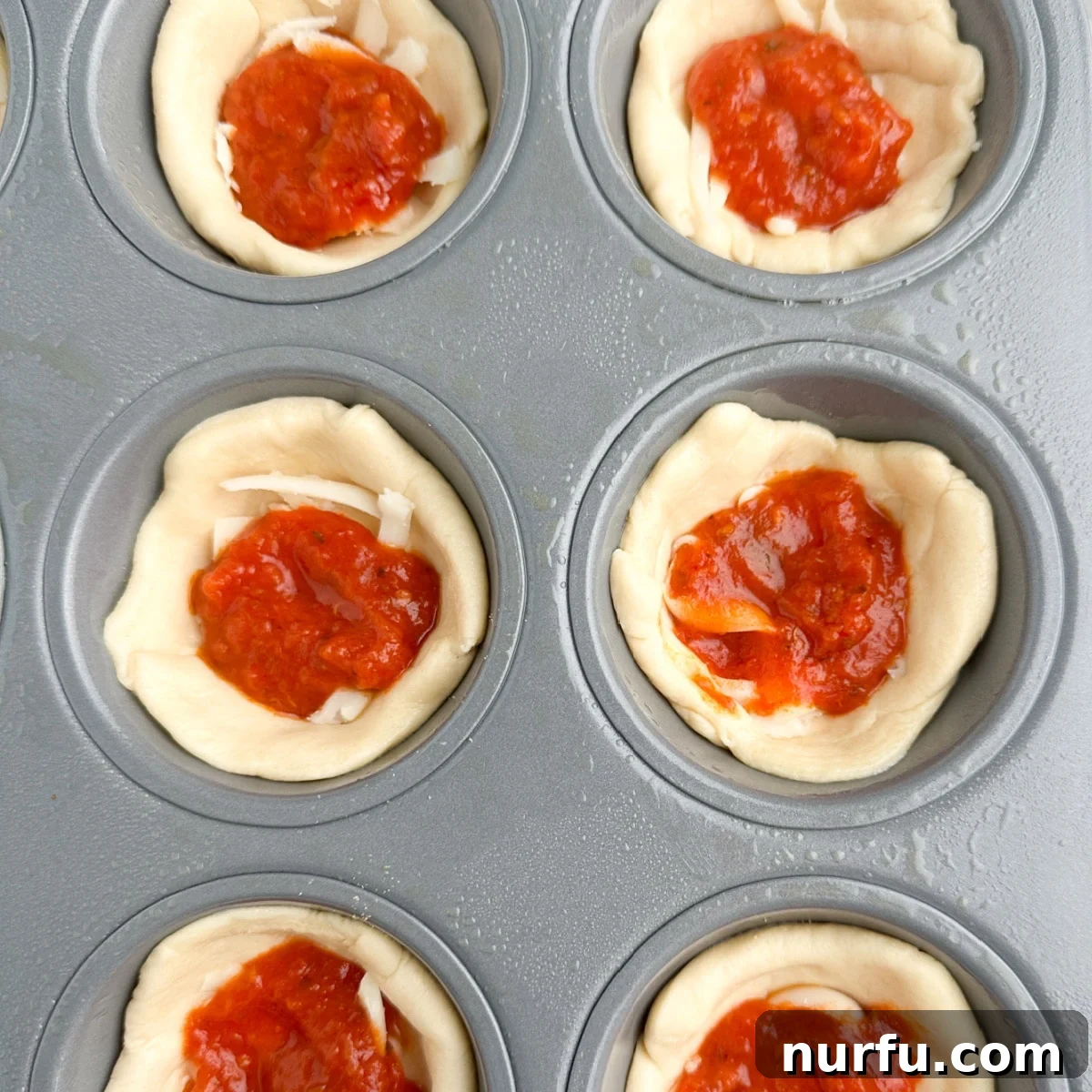 Muffin tin with pizza cupcake bases, now topped with a spoonful of rich red pizza sauce over the cheese layer.