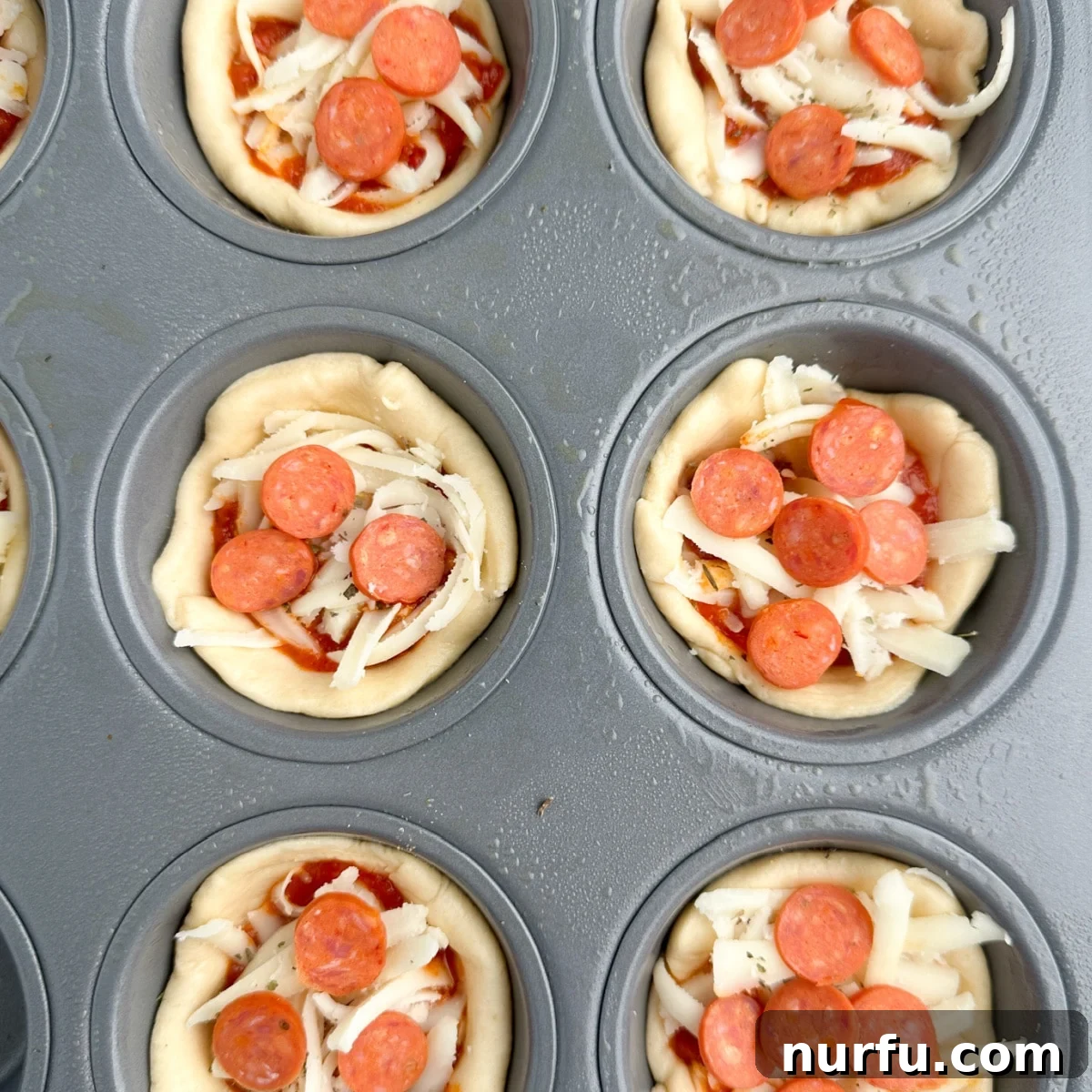 Uncooked pizza cupcakes in a muffin tin, fully assembled with crescent dough, cheese, sauce, Italian seasoning, and mini pepperoni toppings, ready for baking.