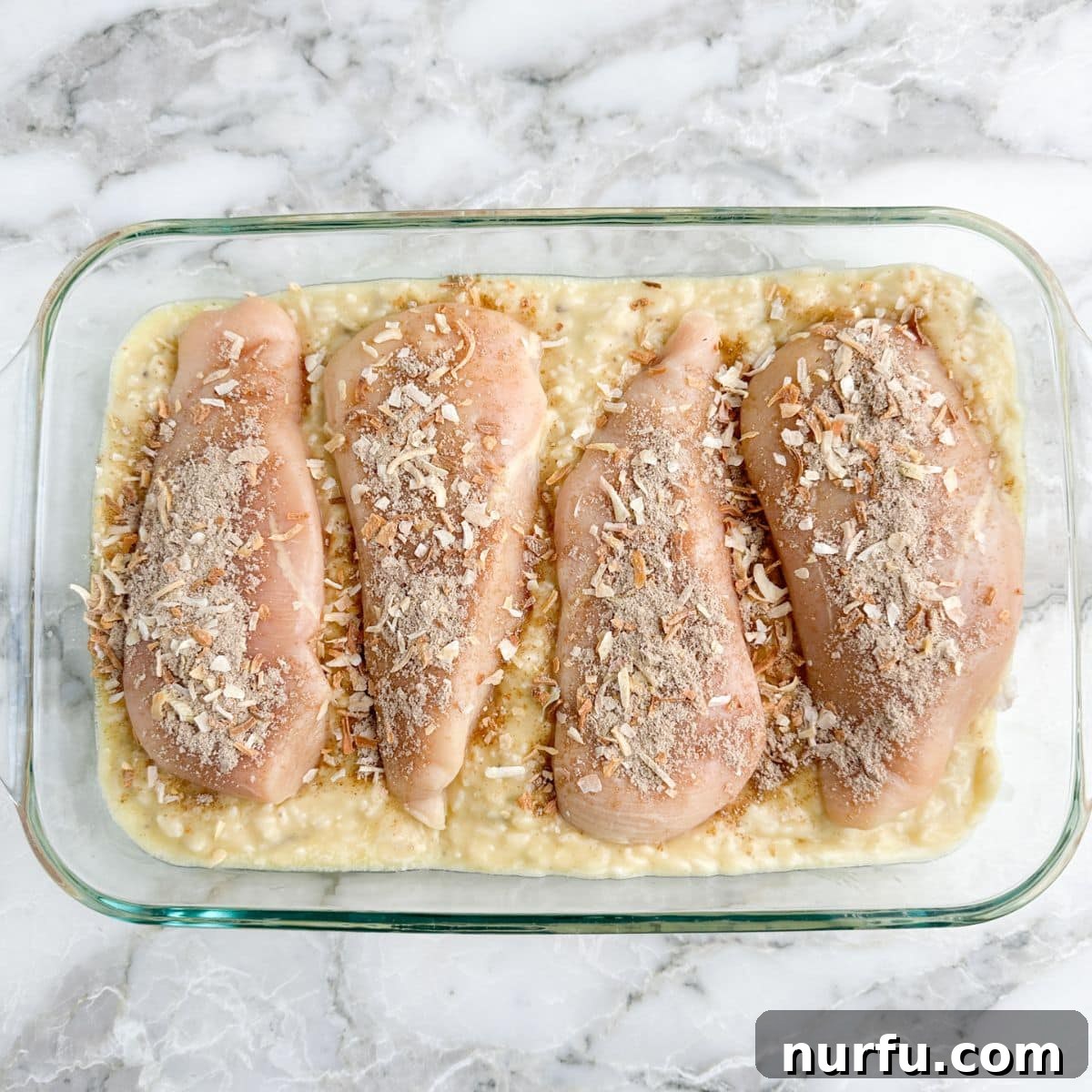 The Chicken That Time Forgot 6 A close-up view of the baking dish, showcasing the creamy rice mixture spread evenly at the bottom, topped with raw chicken breasts ready for seasoning and baking.