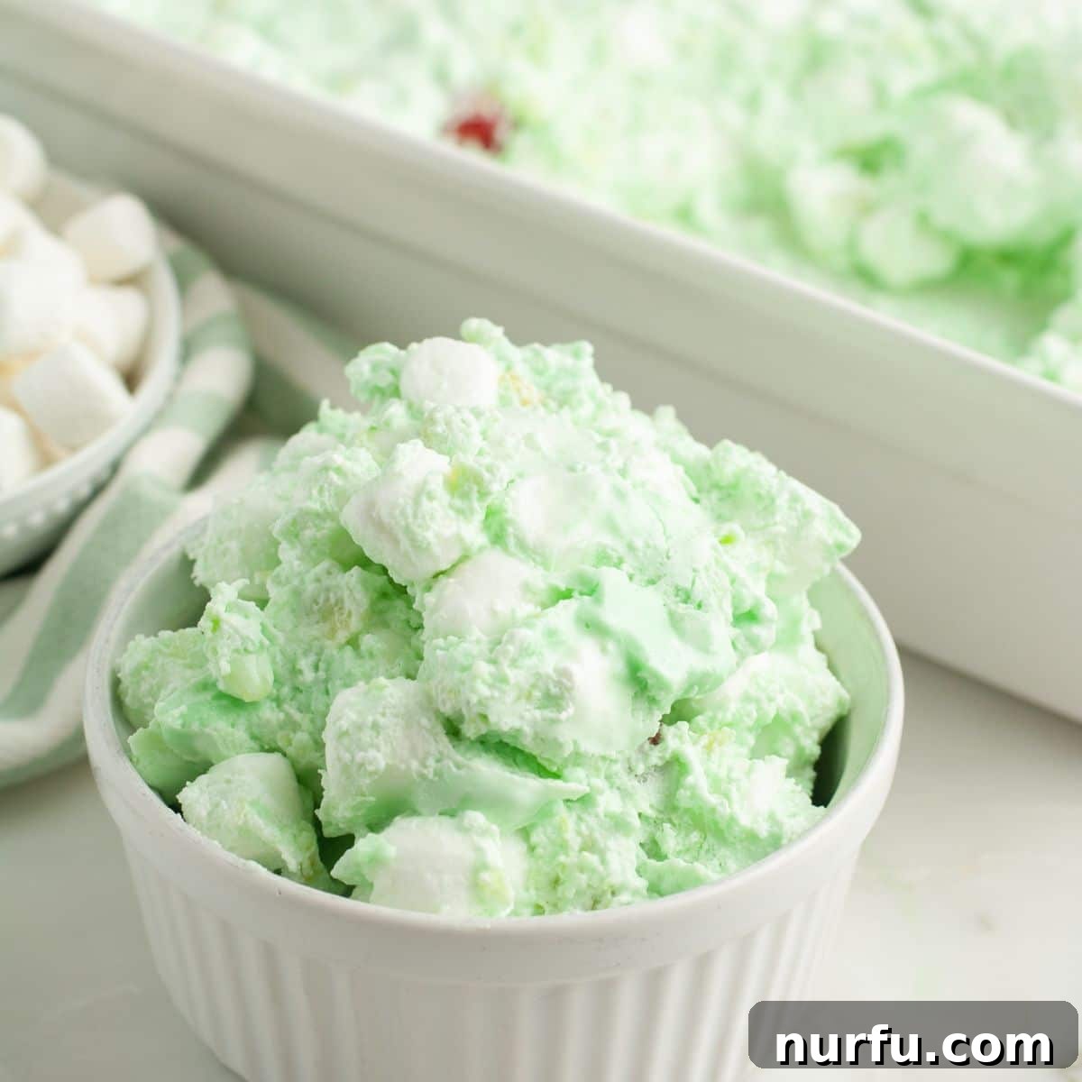 Sea Mist Salad 2 A bowl of creamy, green sea foam salad topped with miniature marshmallows and visible crushed pineapple pieces, served in a decorative white bowl on a light background.