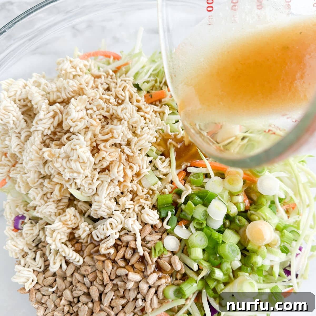 Crunchy Ramen Broccoli Salad 6 Bowl of ramen noodles, sunflower kernels, and broccoli slaw with dressing being poured on it.