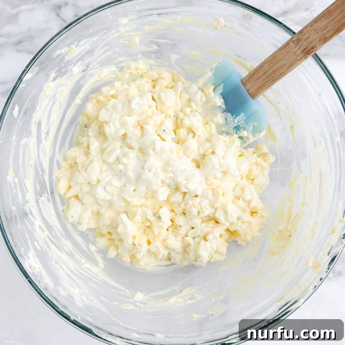 Savory Whipped Egg Dip 5 A bowl showcasing the creamy deviled egg dip mixture with finely chopped hard-boiled eggs gently folded in, being stirred with a spatula before garnishing.