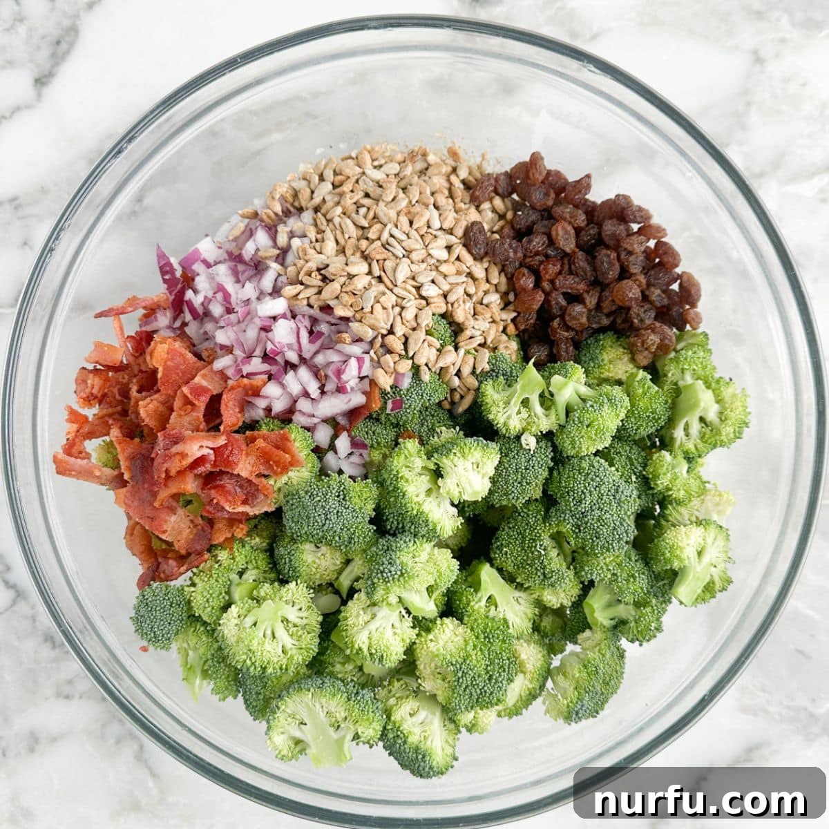 Vibrant Broccoli Raisin Bliss 4 A large mixing bowl containing raw broccoli florets, plump raisins, crumbled crispy bacon, diced red onion, and crunchy sunflower seeds, ready to be tossed with dressing.