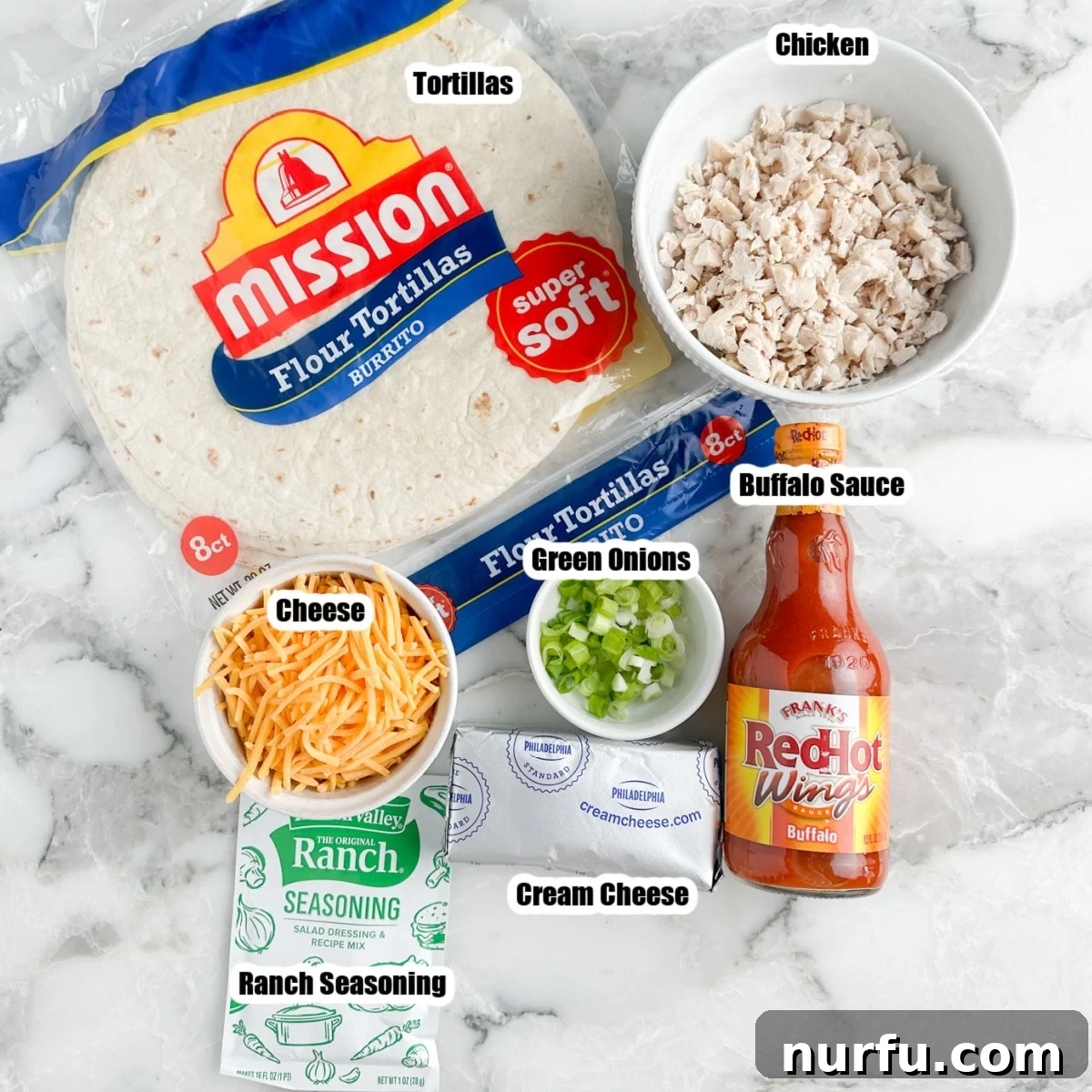 A flat lay photograph showcasing all the ingredients for Buffalo Chicken Pinwheels: flour tortillas, cooked chicken, cream cheese, shredded cheddar cheese, buffalo wing sauce, ranch seasoning, and chopped green onions.
