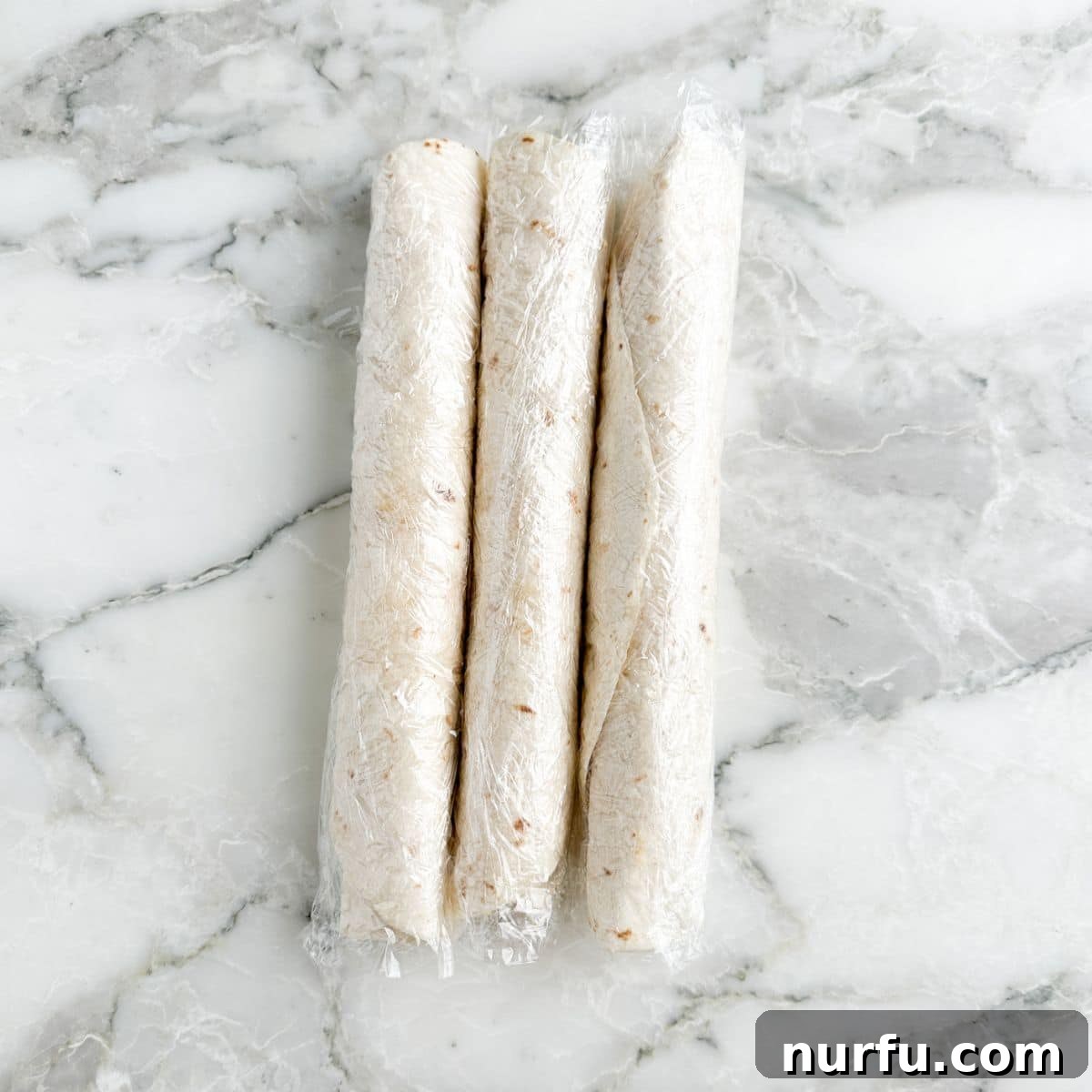 Three tightly rolled tortillas, each individually wrapped in plastic wrap, chilling in the refrigerator.