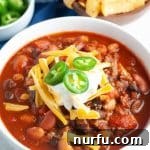A bowl of robust three bean chili, garnished with a dollop of sour cream and fresh jalapeño slices.