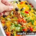 A hand holding a tortilla chip, scooping up a generous amount of layered ground beef taco dip.
