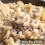 Wooden spoon scooping macaroni noodles and ground beef from a skillet, showcasing a creamy Alfredo sauce.