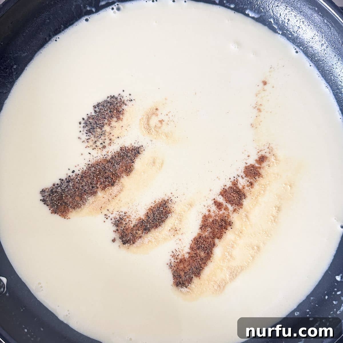Creamy Parmesan Alfredo Spaghetti 5 A whisk stirring a creamy Alfredo sauce in a skillet, with a sprinkle of seasonings like nutmeg, garlic powder, salt, and pepper visible.
