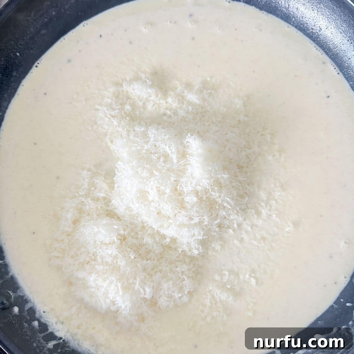 Creamy Parmesan Alfredo Spaghetti 6 A close-up of a skillet filled with creamy Alfredo sauce, with a mound of freshly grated Parmesan cheese being whisked in.