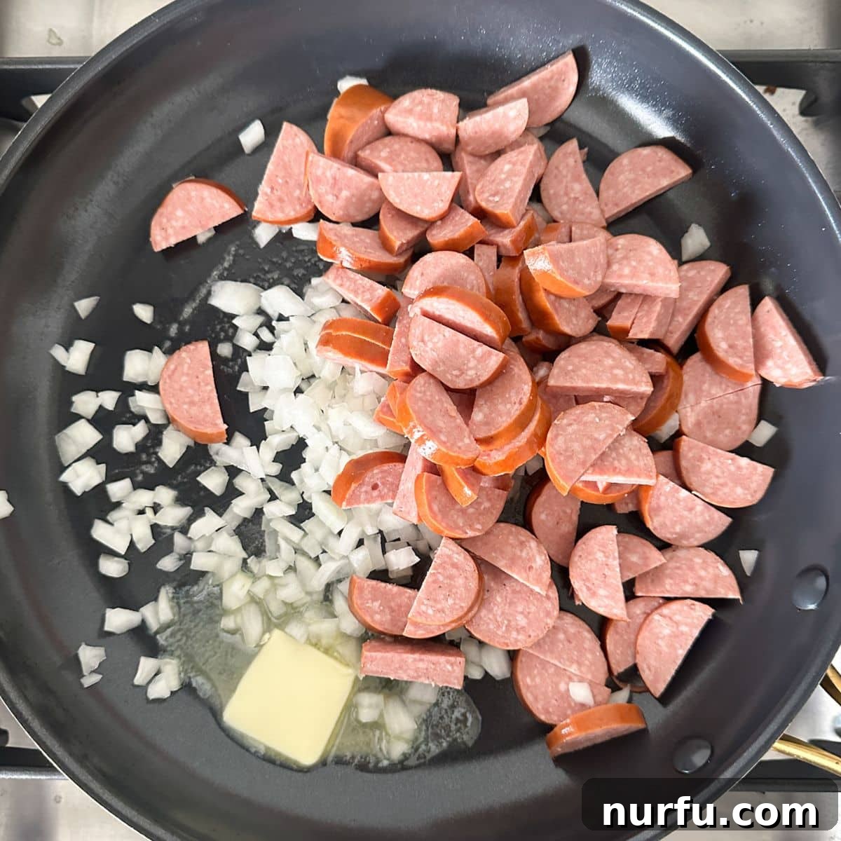 Hearty Kielbasa Pasta 4 A large skillet on a stovetop containing browned slices of kielbasa and softened diced onions, sizzling in butter, with a spatula for stirring.