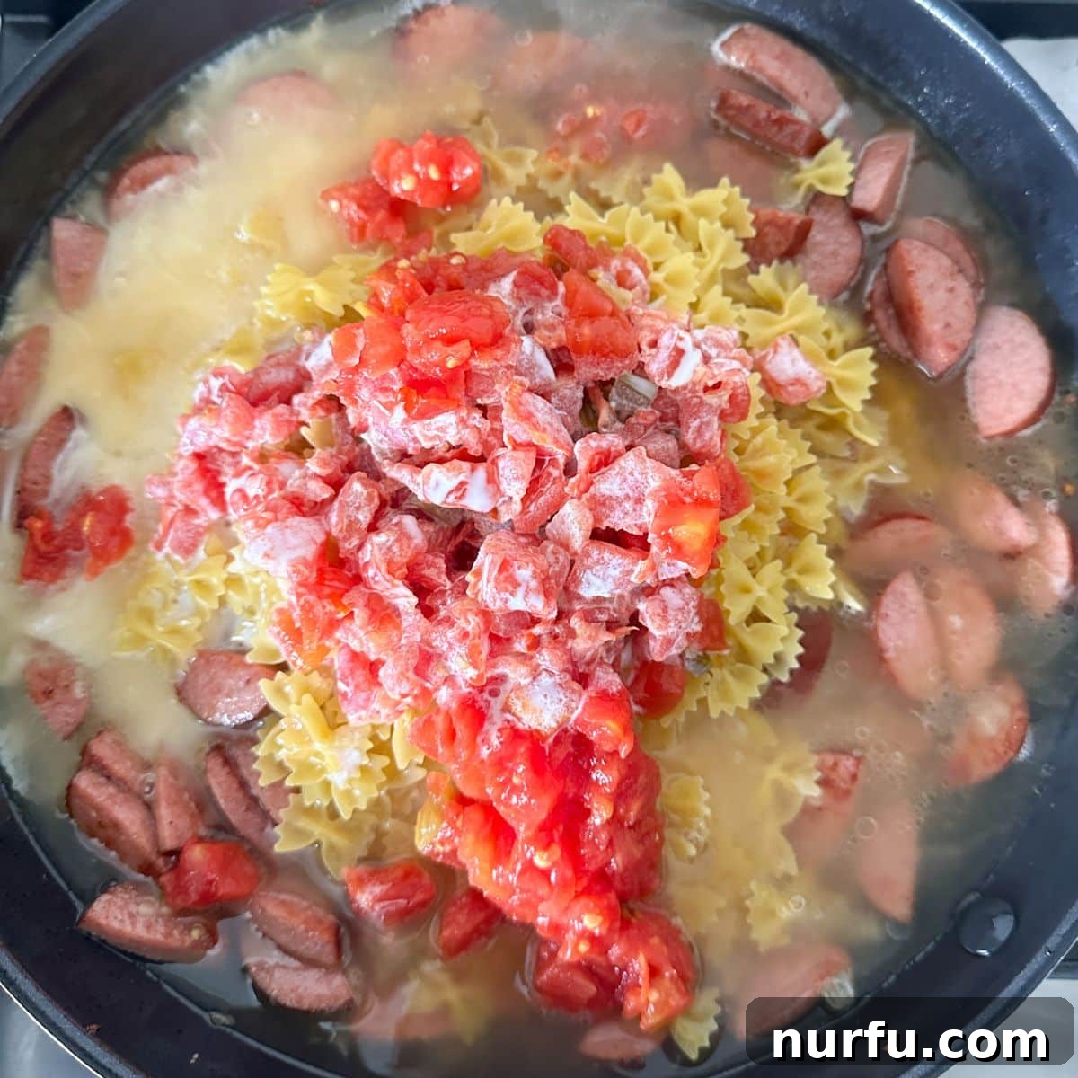 Hearty Kielbasa Pasta 5 A skillet filled with browned sausage and onions, now with dry pasta, Rotel tomatoes, and chicken broth added, being stirred to combine before simmering.
