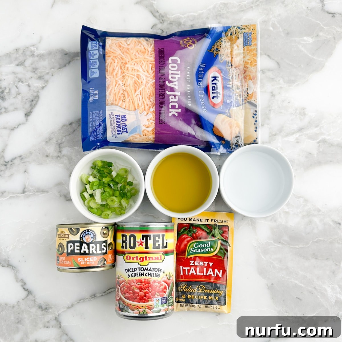 Poolside Refresh 3 A carefully arranged photo of all the ingredients for poolside dip: a bag of shredded Colby Jack cheese, a bowl of diced green onions, a bottle of extra virgin olive oil, white vinegar, a can of sliced black olives, a can of ROTEL diced tomatoes, and a packet of Zesty Italian seasoning mix.