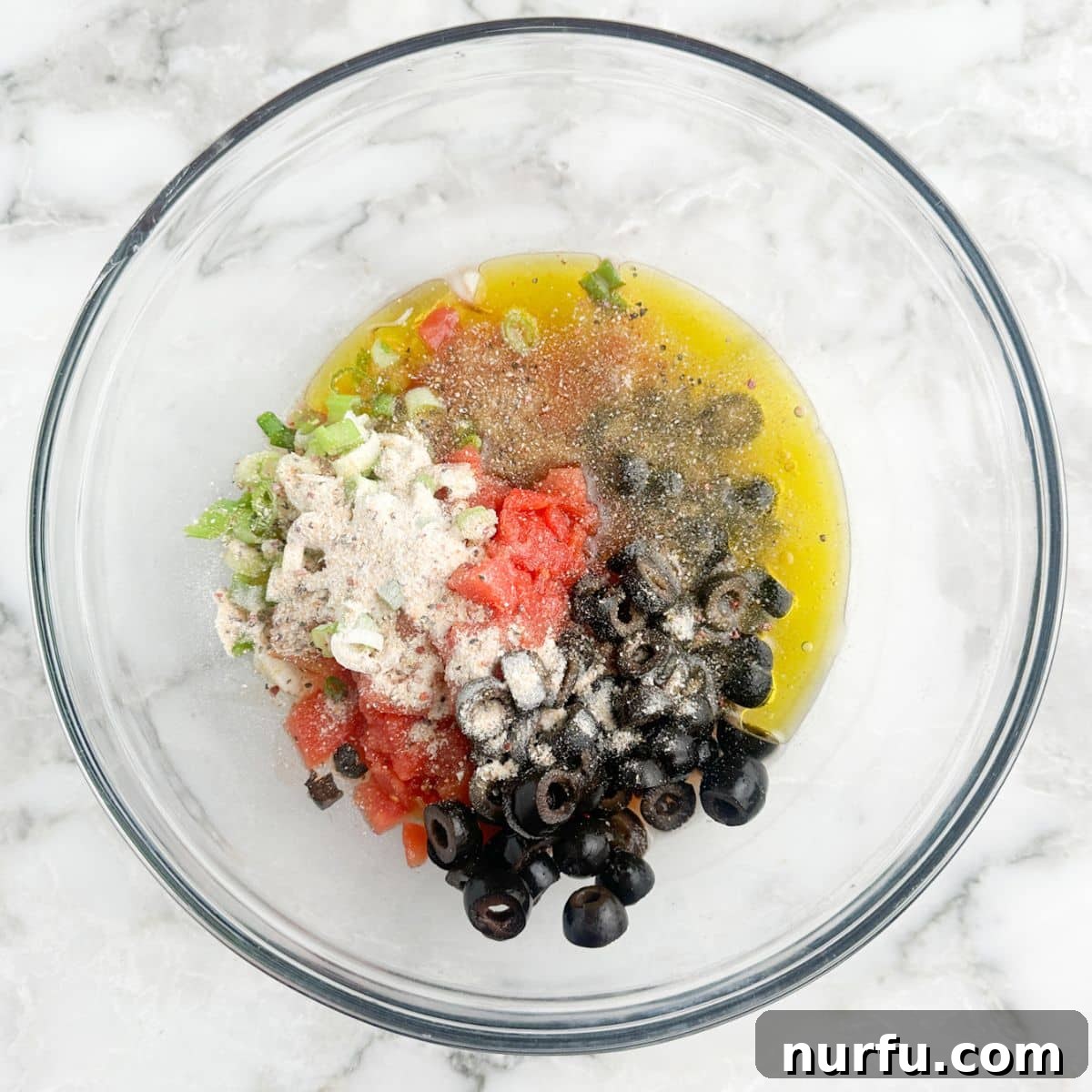 Poolside Refresh 4 A large mixing bowl containing a colorful array of ingredients for poolside dip: drained black olives, diced Rotel tomatoes, zesty Italian seasoning, white vinegar, and a drizzle of extra virgin olive oil.