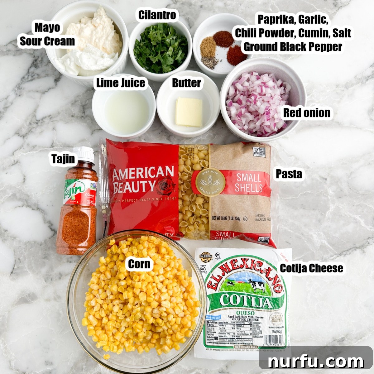 Elote Fiesta Pasta 3 A vibrant flat lay of elote pasta salad ingredients including pasta, a bowl of corn, crumbled cotija cheese, chopped red onions, Tajin seasoning, mayonnaise, and fresh cilantro, alongside various spices.