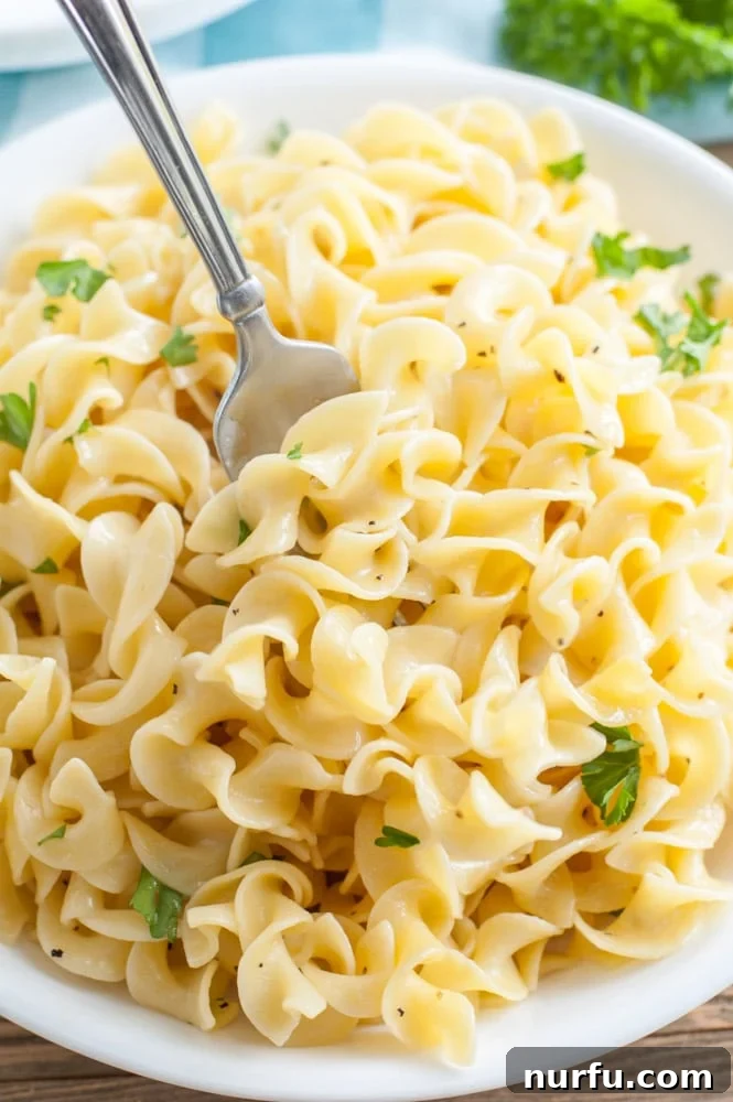 Buttered Noodles A white bowl filled with golden buttered noodles, garnished with fresh parsley, with a fork resting inside.