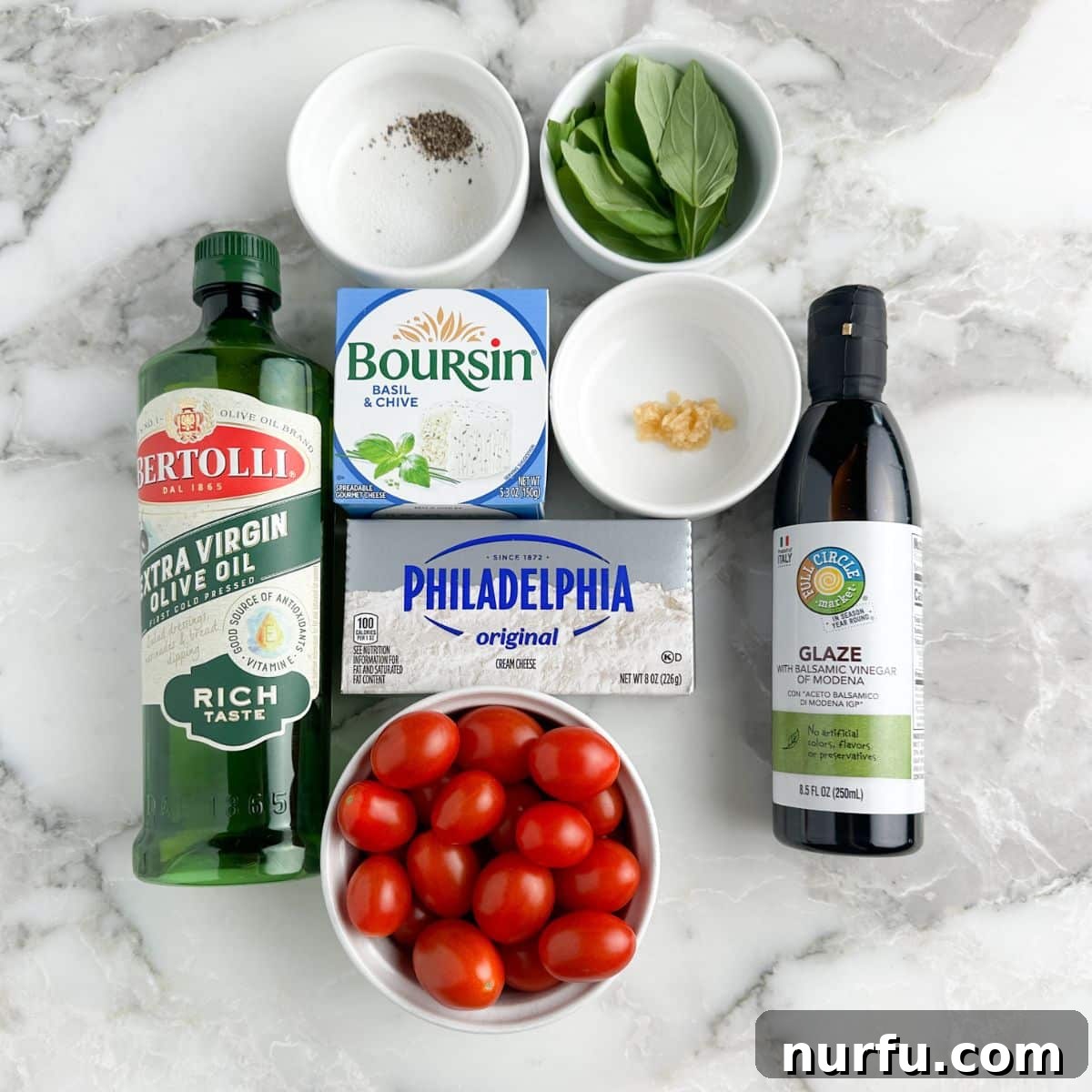 Sun-Kissed Bruschetta Topping 3 A flat lay photograph displays the key ingredients for bruschetta dip: a bottle of extra virgin olive oil, balsamic glaze, fresh red tomatoes, a block of cream cheese, a round of Boursin cheese, and vibrant green basil leaves, all arranged neatly on a surface.