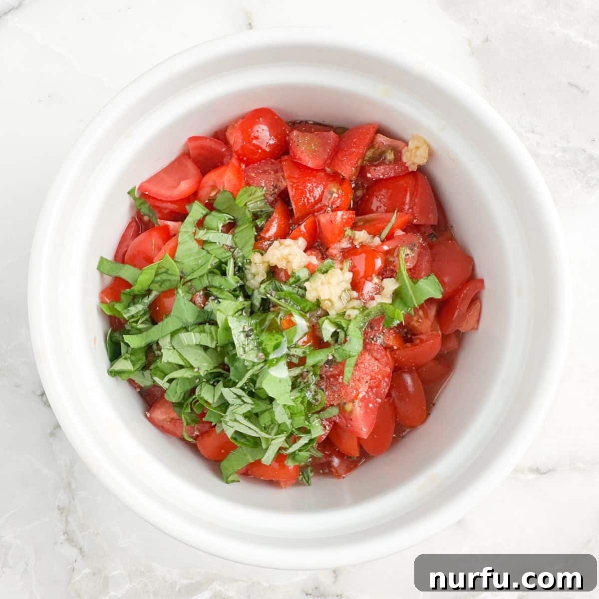 Sun-Kissed Bruschetta Topping 6 A white bowl holds a vibrant mixture of finely diced red tomatoes and fresh green basil leaves, seasoned and ready to be used as the bruschetta topping.