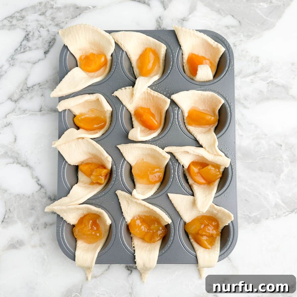 Flaky Peach Cheesecake Delights 4 Muffin tin with crescent roll dough pressed into cups, each filled with peach pie filling.