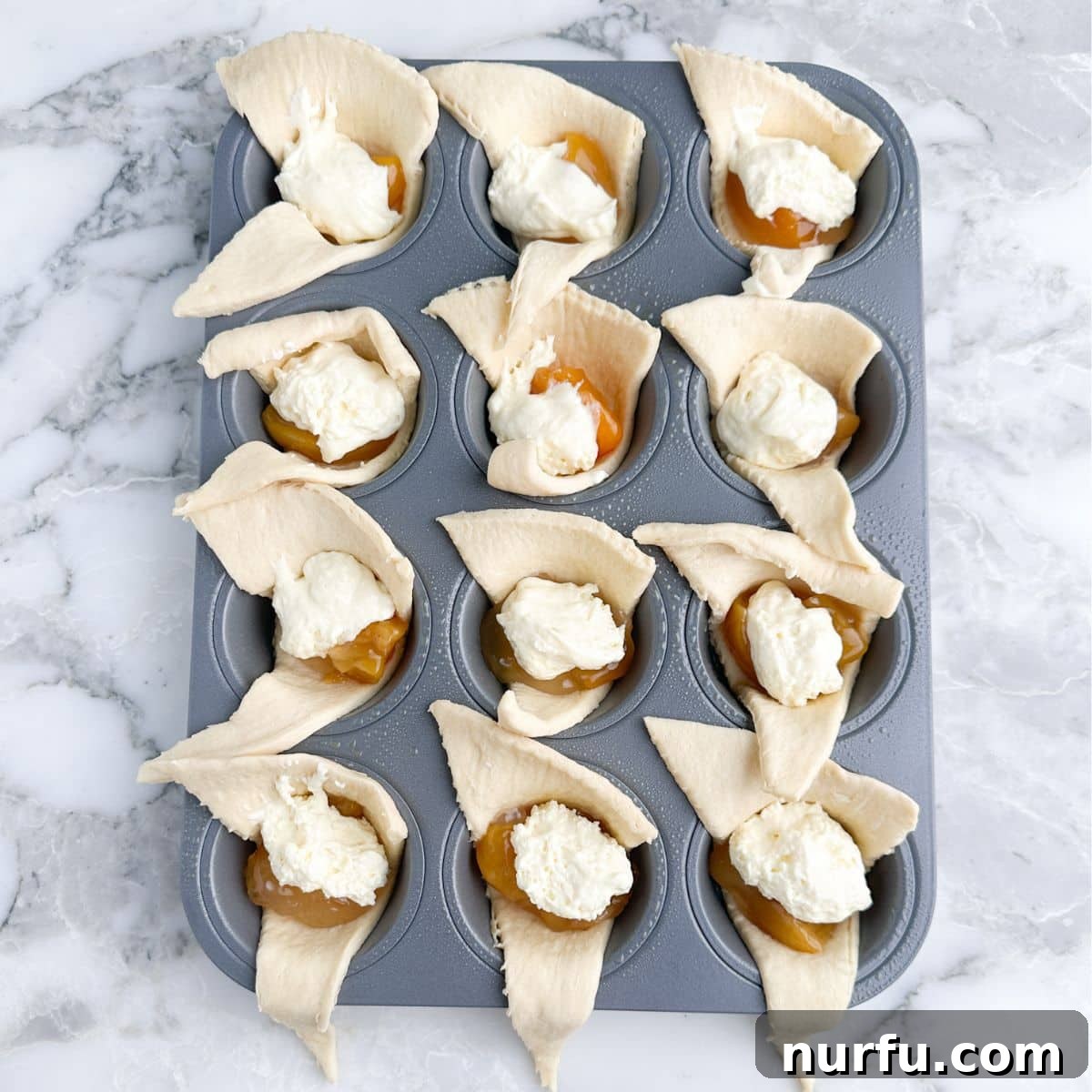 Flaky Peach Cheesecake Delights 5 Muffin tin with uncooked crescent rolls, peach pie filling, and a dollop of cheesecake filling on top.