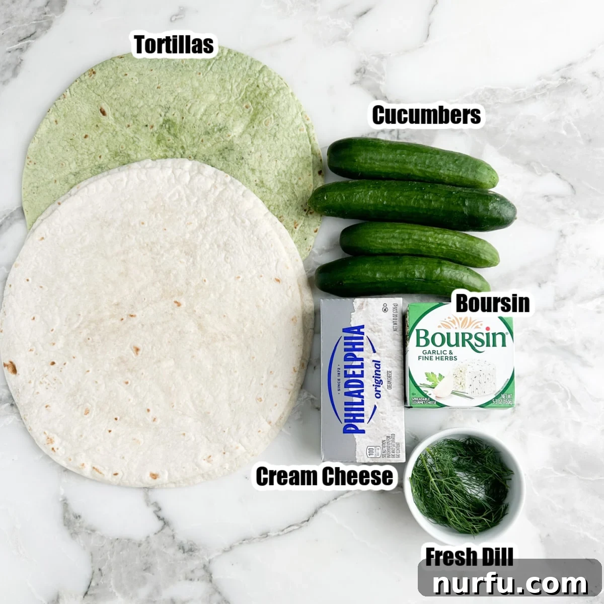 A flat lay photograph showcasing the key ingredients: flour tortillas, fresh cucumbers, cream cheese, Boursin cheese, and a sprig of fresh dill.