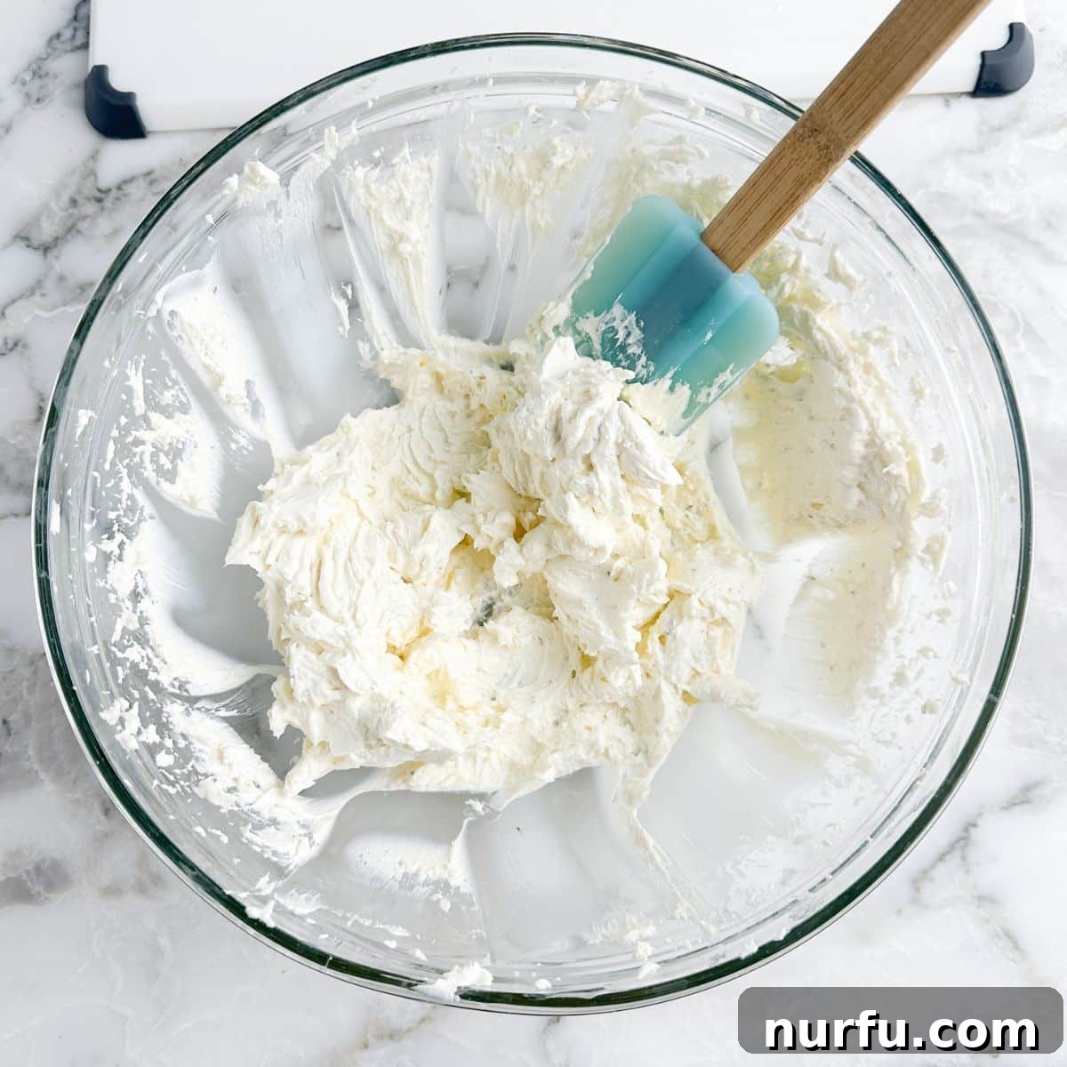 A mixing bowl containing softened cream cheese and Boursin cheese, with a spatula ready for creaming.