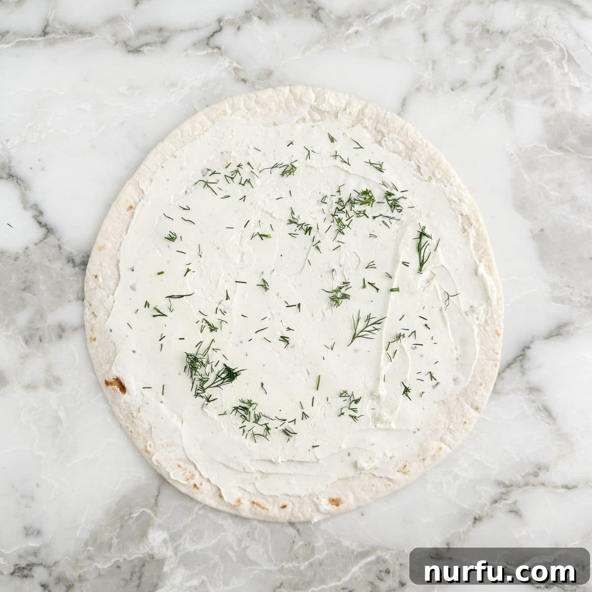A flat tortilla generously spread with the cream cheese mixture, ready for the next layer.