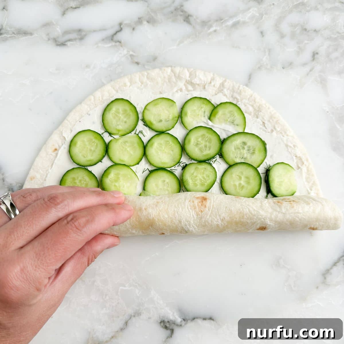 A tortilla with cream cheese and sliced cucumbers being tightly rolled up into a cylinder.