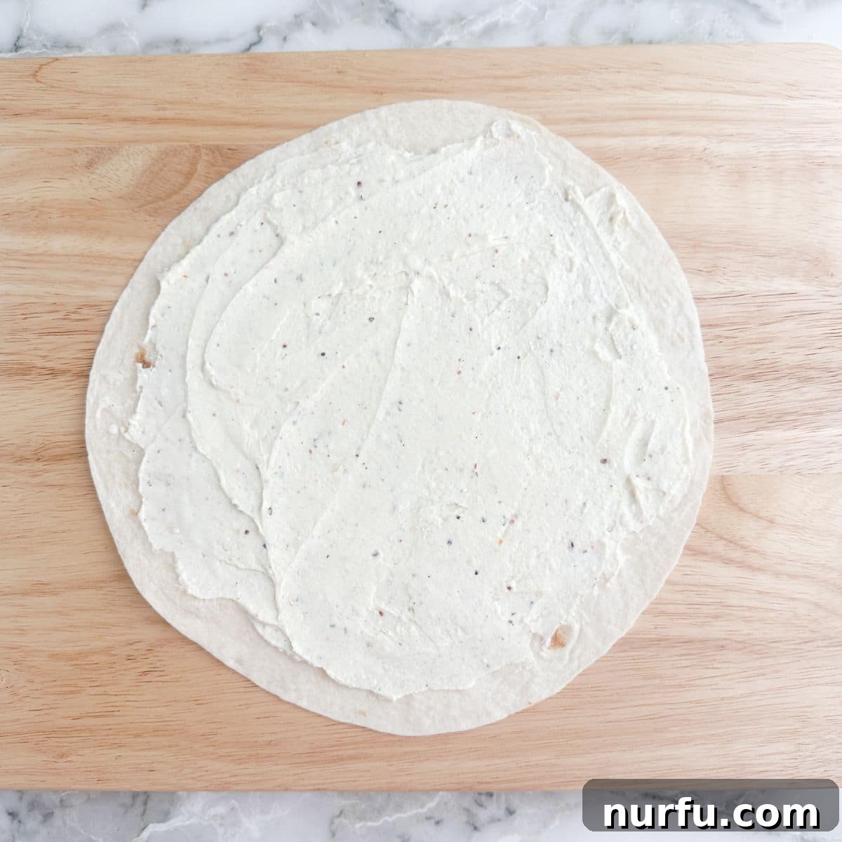 Creamy BLT Roll-Ups 4 A large flour tortilla evenly covered with a creamy, pale yellow spread from edge to edge, ready for toppings.
