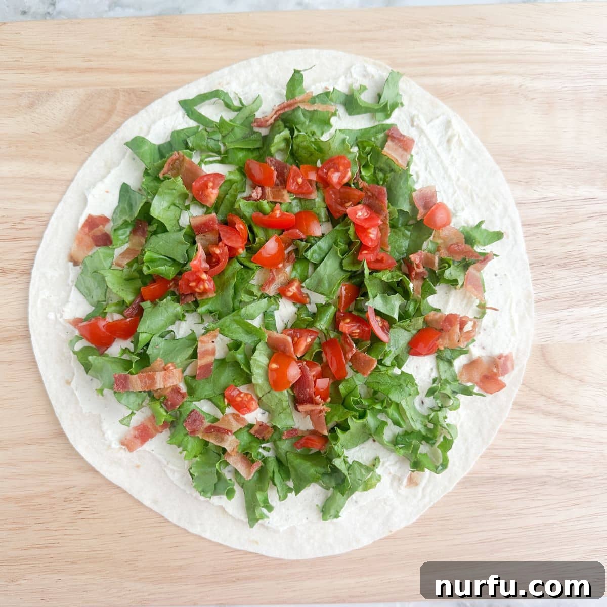 Creamy BLT Roll-Ups 5 A tortilla with creamy spread, generously topped with finely chopped lettuce, crumbled crispy bacon, and diced red tomatoes, preparing for rolling.