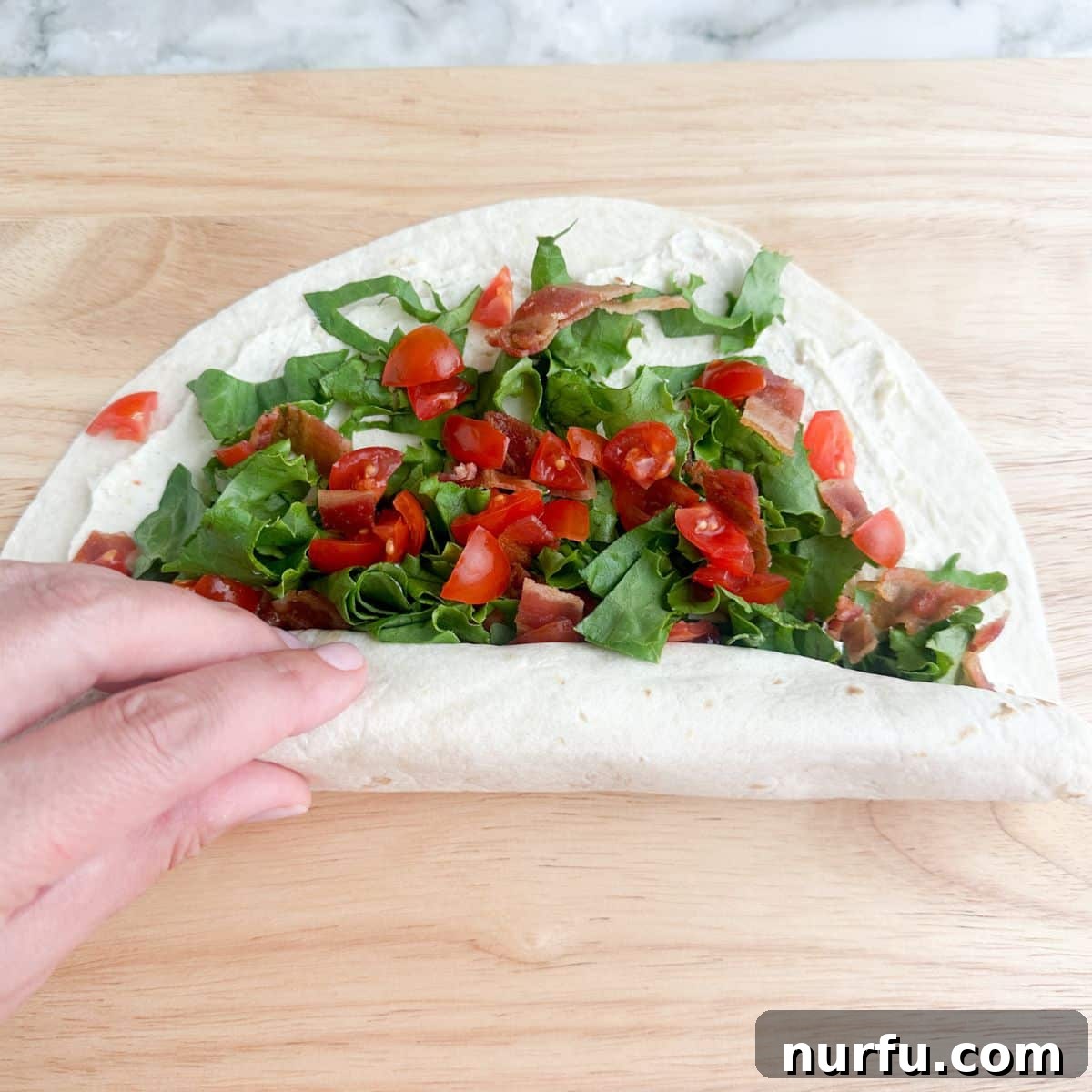 Creamy BLT Roll-Ups 6 A tortilla, fully topped with BLT ingredients, being tightly rolled from one end to the other to form a compact wrap.