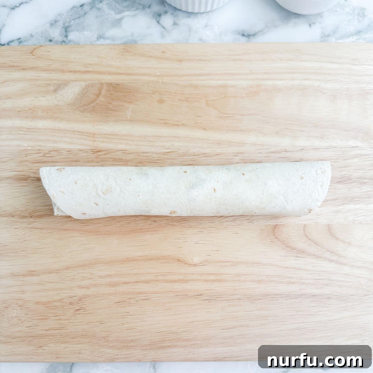 Creamy BLT Roll-Ups 7 A tightly rolled tortilla wrap, covered in plastic wrap, resting on a wooden cutting board, ready for chilling and slicing into pinwheels.