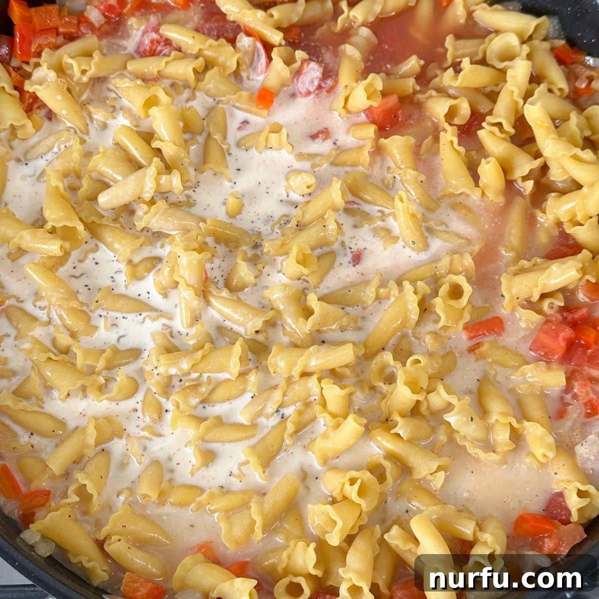 Uncooked pasta submerged in a creamy, tomato-based sauce within a skillet, covered and ready to cook until tender.