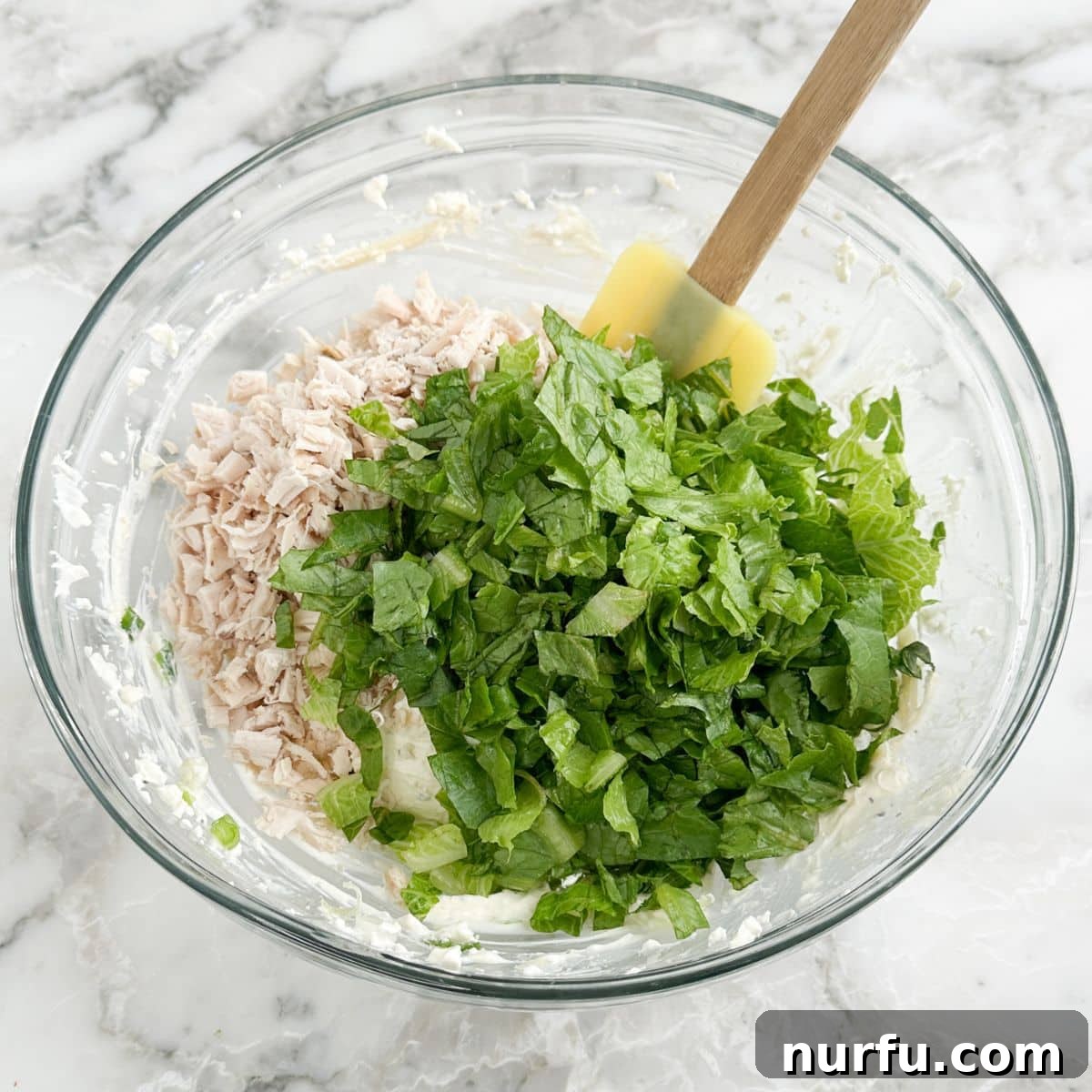 A large bowl containing shredded chicken, finely chopped romaine lettuce, and the creamy Caesar dressing mixture, being stirred together.